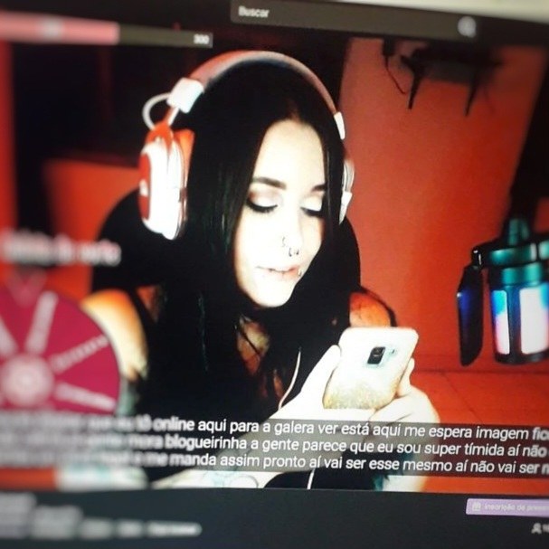 Registro de uma live: a streamer de headphone olha o celular, com legendas automáticas do Stream-CC na parte de baixo da tela